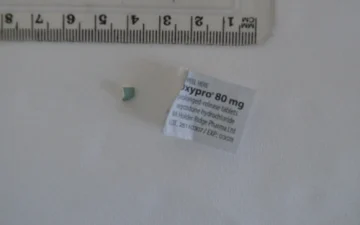 Oxycodon-Tablettenstück neben dem Etikett einer Oxypro 80 mg Retardpackung